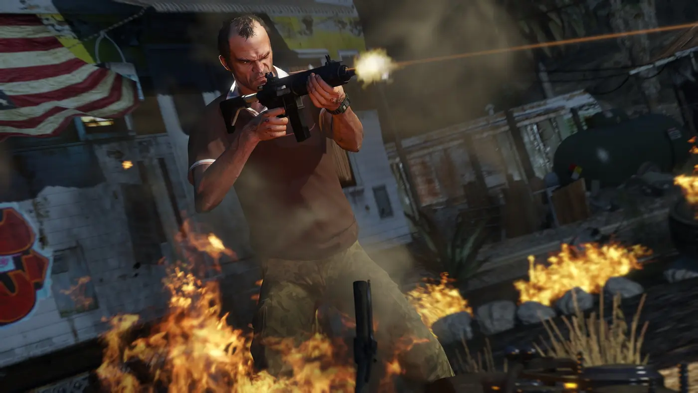 Trevor Philips firing a submachine gun amid flames and smoke in a run-down yard, an American flag hanging on a wall behind him