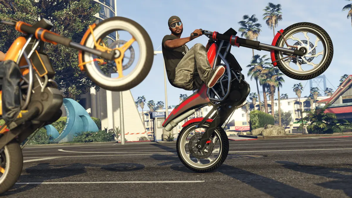 A rider in a cap and sunglasses pops a wheelie on a red motorcycle in a palm-lined parking lot, with a second bike in the foreground