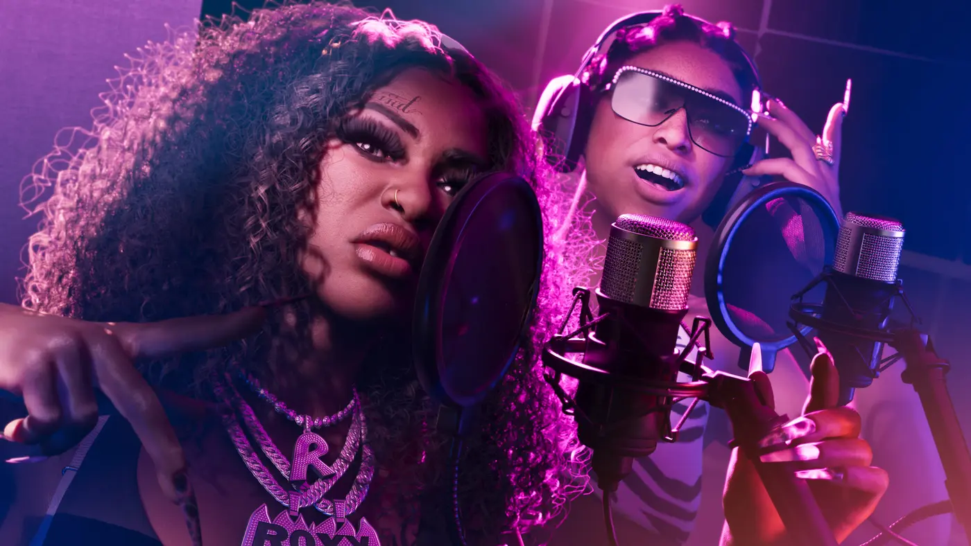 Two female GTA VI artists record in the studio, leaning toward professional mics fitted with pop filters, headphones on, glittery glasses and a ROXY pendant, bathed in pink and purple neon light