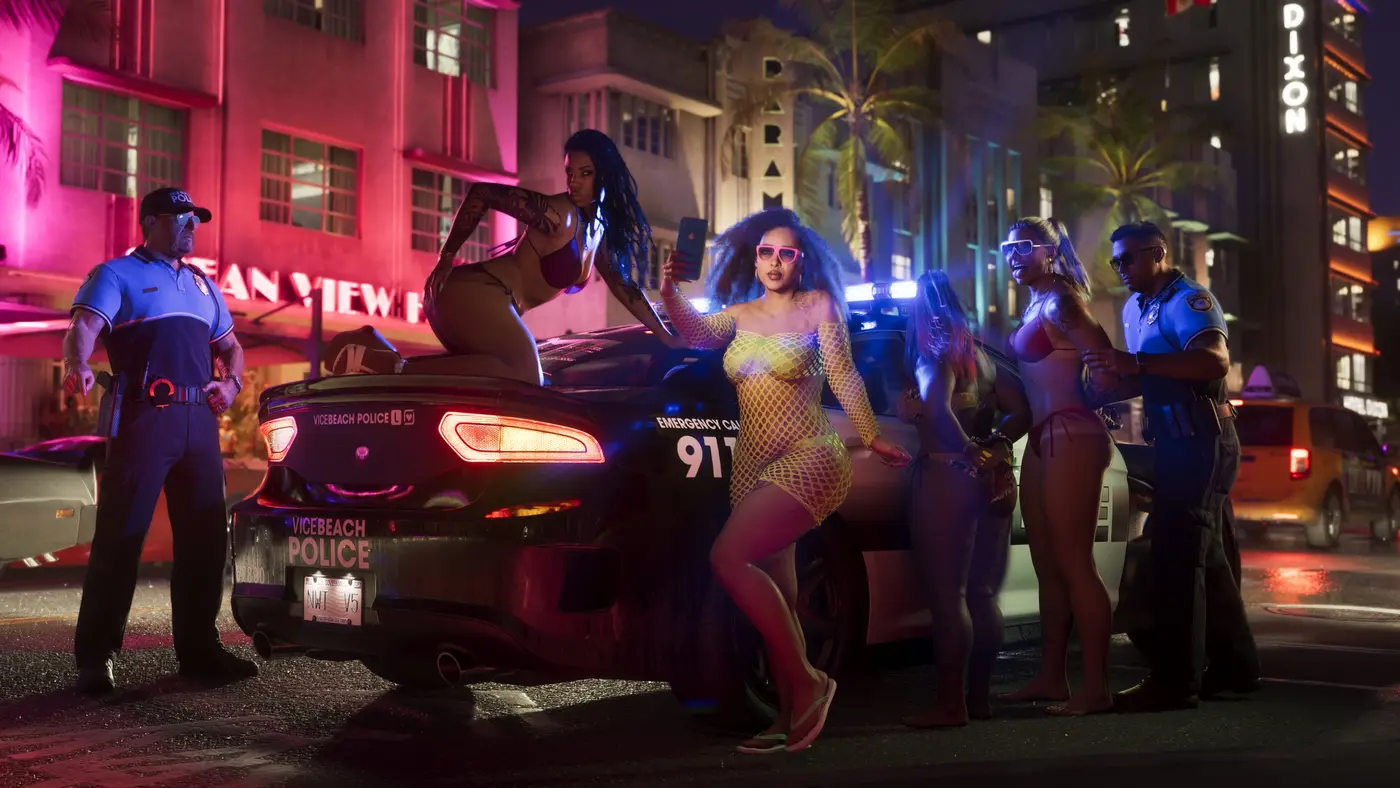 A Vice City street at night in GTA 6, a group of young women, one taking a smartphone selfie in front of a black police car marked VICEBEACH POLICE and EMERGENCY CALL 911, two officers and pink-neon Art Deco hotels