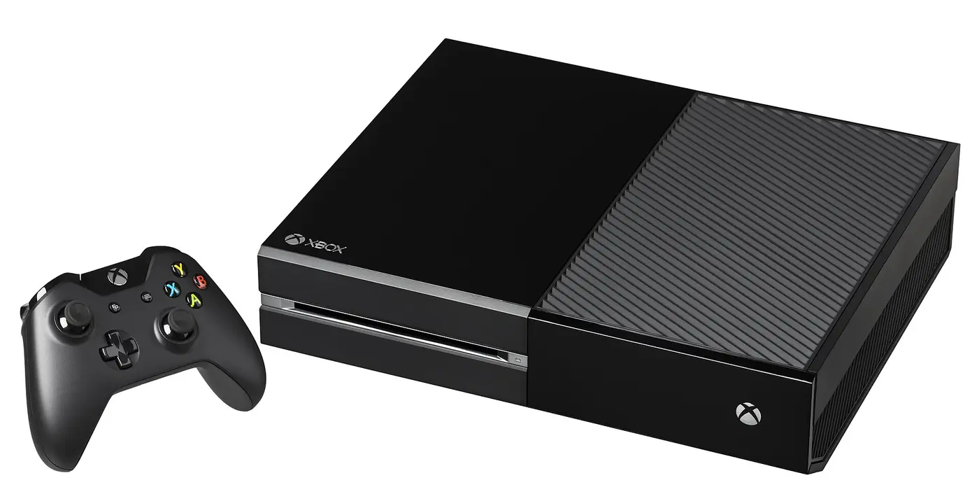A black 2013 Xbox One lying horizontally with its black controller, on a white background